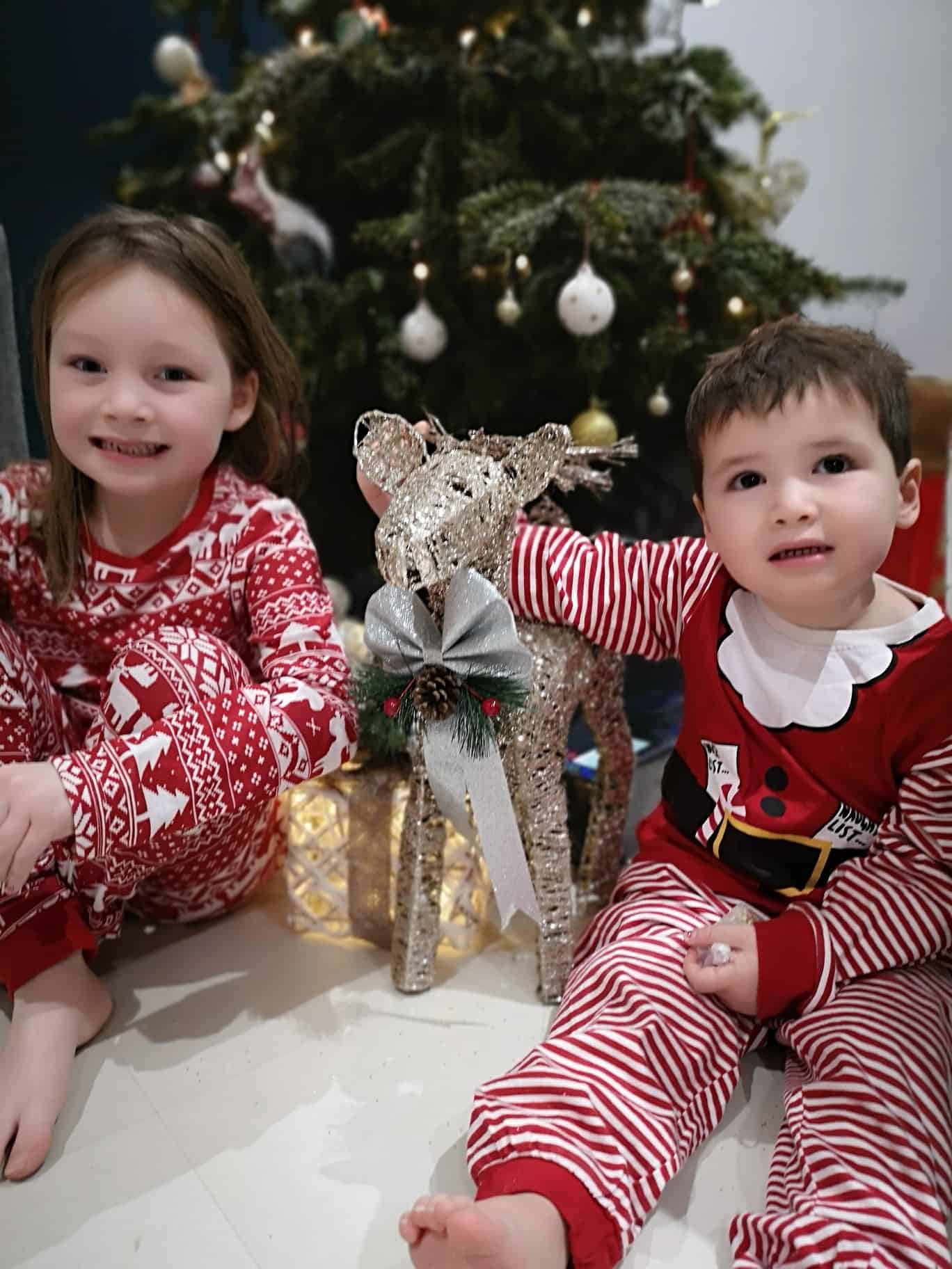 Two children dressed in Christmas clothes sat by Christmas tree